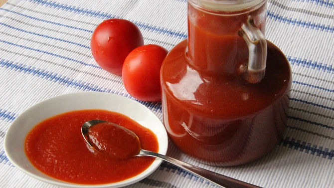 Sklenice domácího kečupu s čerstvými rajčaty na pruhovaném ubrusu.