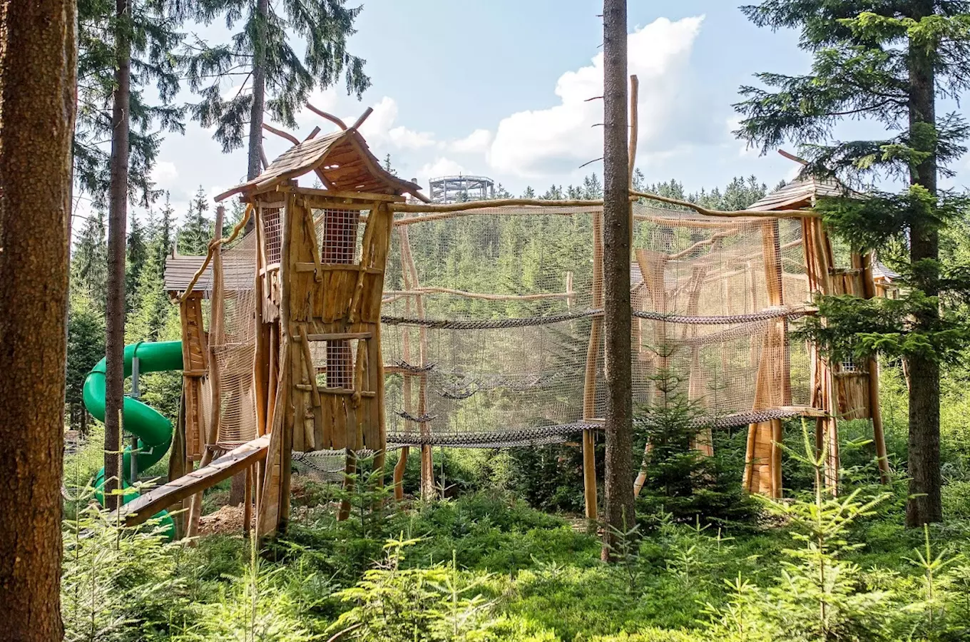 Dřevěné hřiště v lese s houpačkou a skluzavkou.