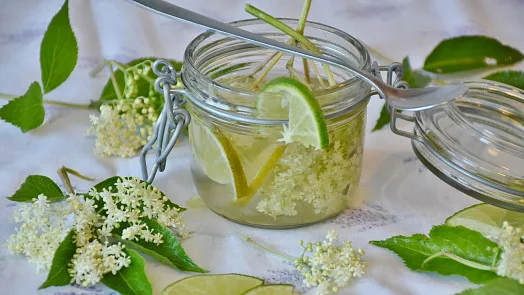 Sklenice s bezovou limonádou, citronem a květy bezu na bílém ubrusu.