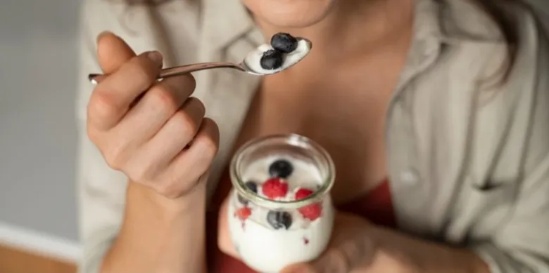 Žena drží lžičku jogurtu s borůvkami nad sklenicí.
