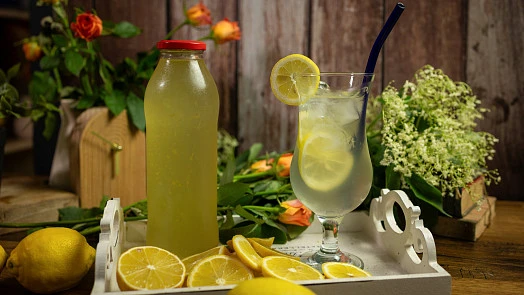 Sklenička limonády s citronem, květinami na dřevěném podnose.