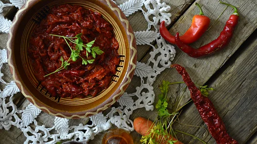 Domácí ajvar v misce s čerstvými paprikami a petrželkou na dřevěném povrchu.