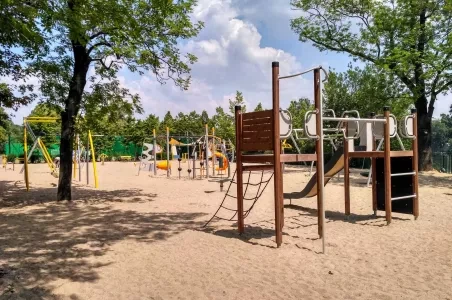 Venkovní dětské hřiště s dřevěnou hrací věží uprostřed lesoparku.